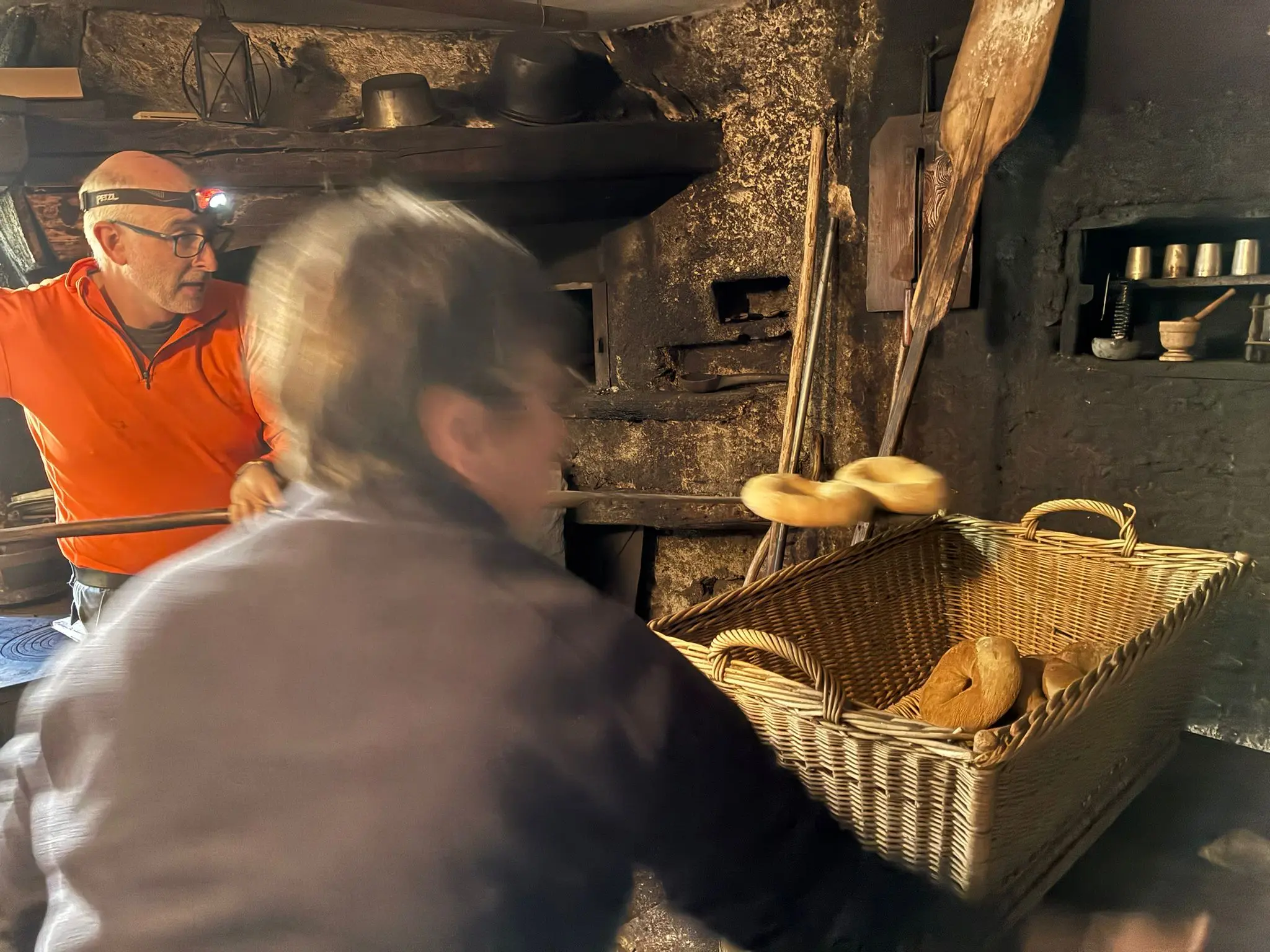 Due persone in un'antica cucina. Un uomo indossa una lampada frontale e si prepara a estrarre pane da un forno. Un altro uomo sta muovendo un cesto con del pane appena cotto. Le pareti sono in pietra, con utensili rustici appesi.