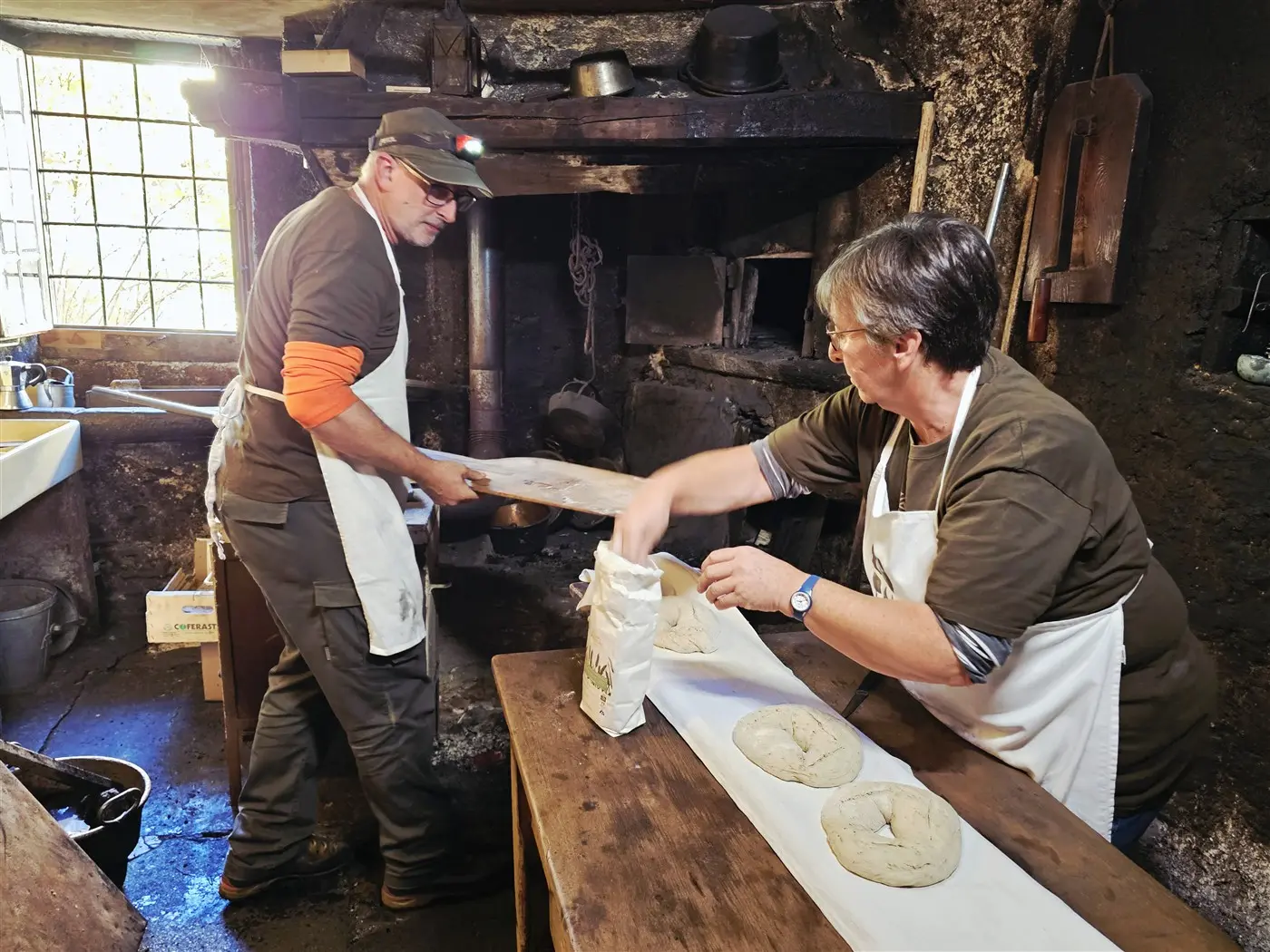 Due persone stanno preparando il pane in una cucina rustica. Un uomo sta sollevando una teglia con il pane mentre una donna stende l'impasto su un tavolo di legno. L'ambiente è caratterizzato da pareti scure e utensili da cucina antichi.