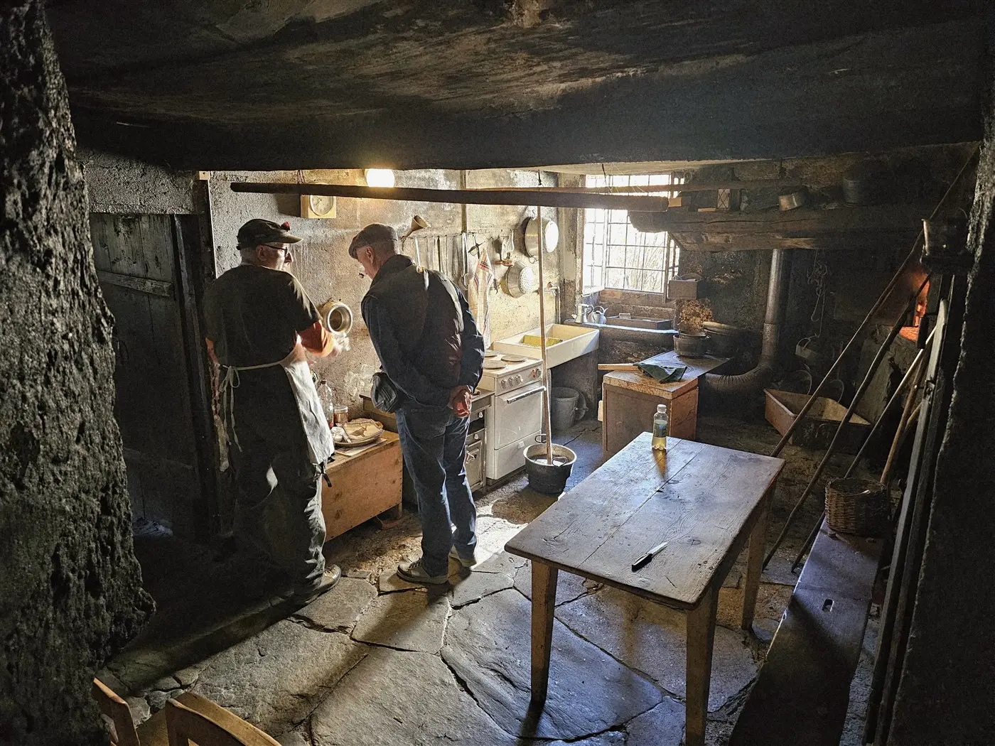 Due uomini nella cucina di un'antica casa, con pareti in pietra e un pavimento in lastre. Uno indossa un grembiule e sta preparando del cibo, mentre l'altro osserva. La luce entra da una finestra, illuminando l'ambiente rustico. Un tavolo in legno è al centro della scena.