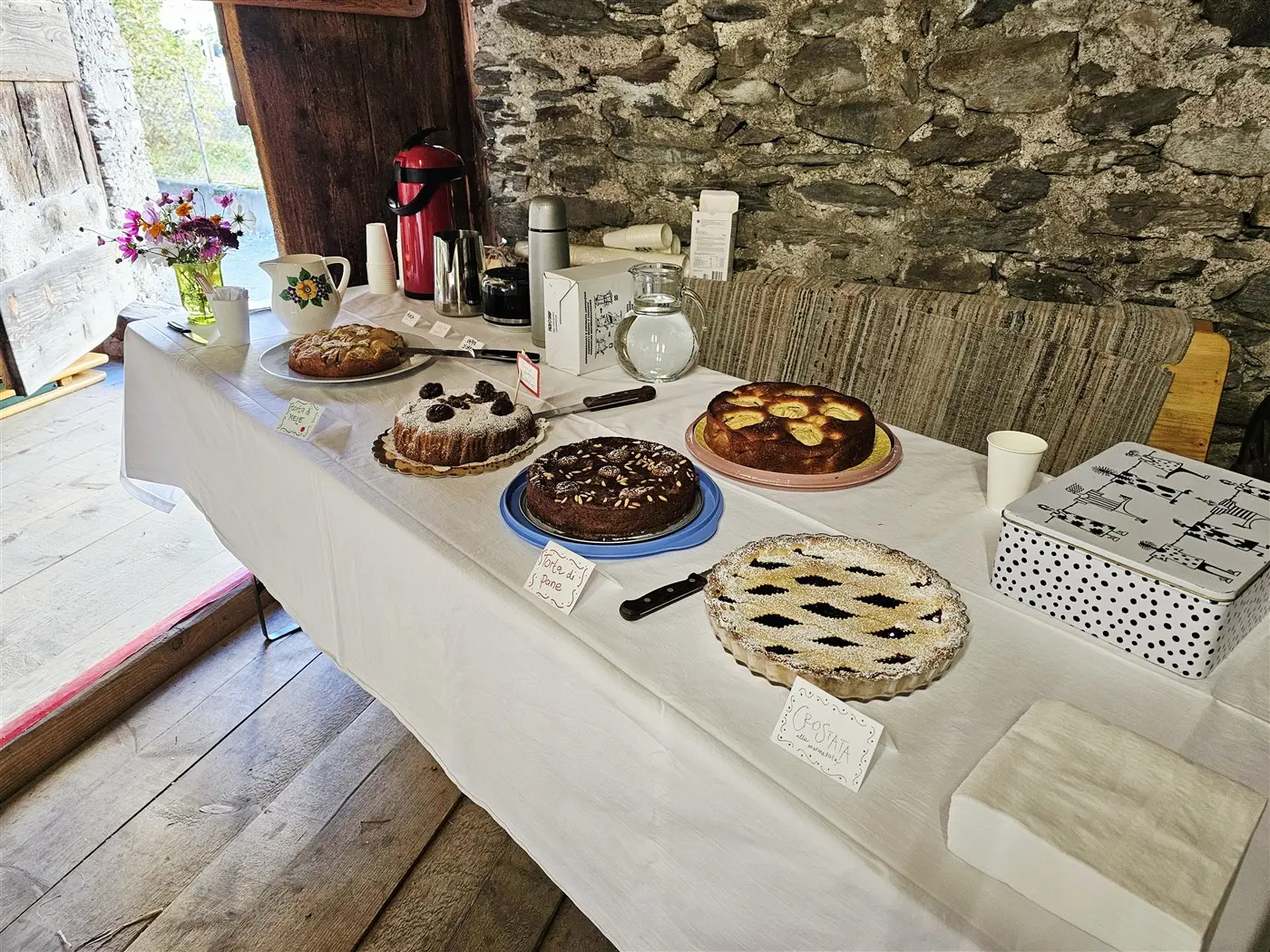 Un tavolo allestito con diverse torte, tra cui dolci al cioccolato e frutta, tutte etichettate. Sullo sfondo si vede un muro di pietra e fiori freschi in un vaso.