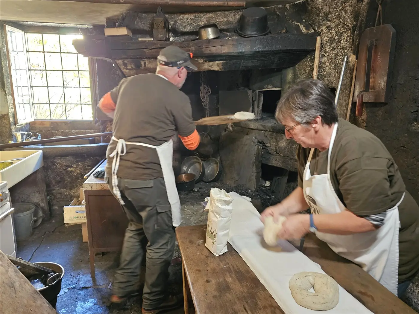 Due persone in un'antica cucina. Un uomo sta inserendo pane nel forno, mentre una donna prepara dell'impasto su un tavolo. L'ambiente è rustico, con attrezzi di cucina e recipienti visibili.