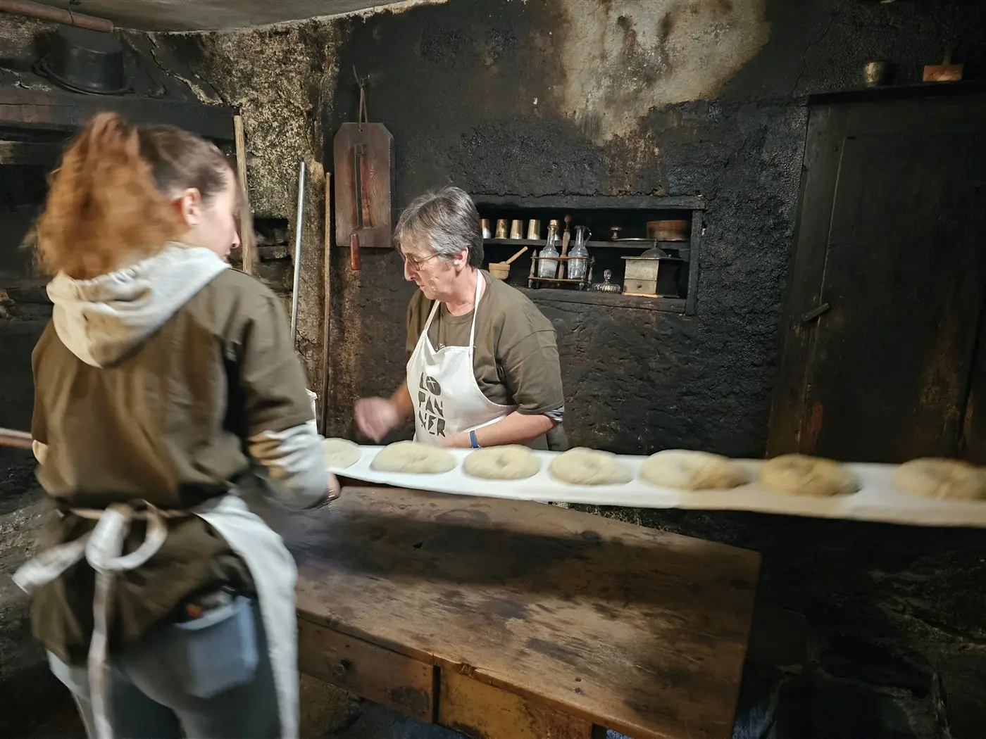 Due donne in un ambiente di panificazione tradizionale, una delle quali prepara impasti su una tavola di legno mentre l'altra osserva. La stanza è caratterizzata da pareti nere e attrezzi da cucina appesi.