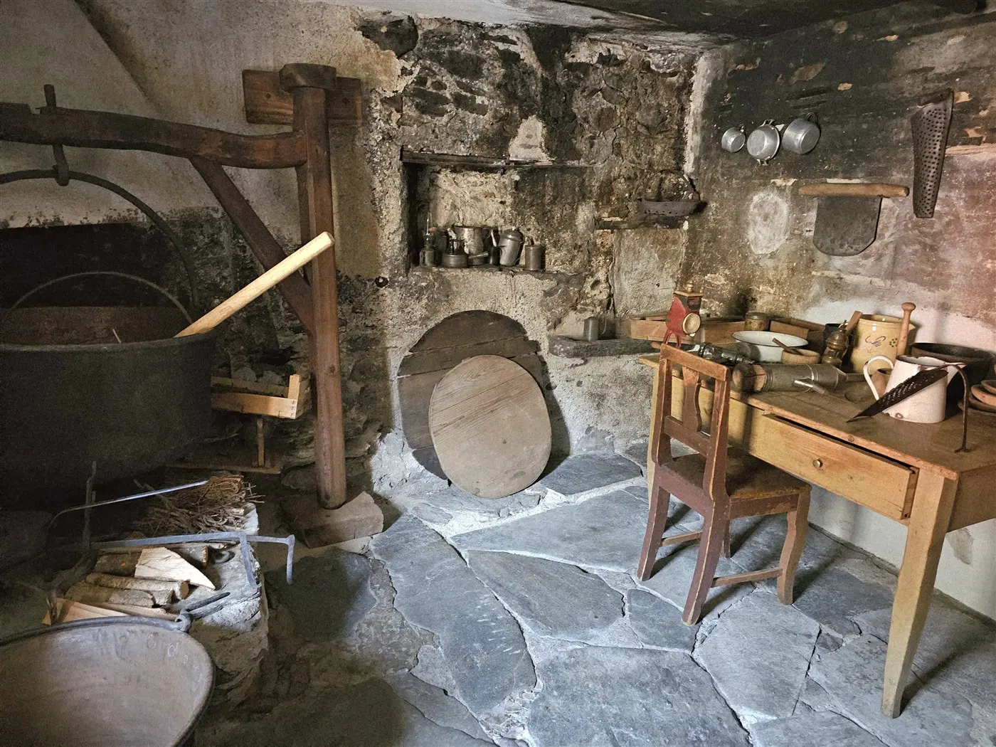 Una cucina rustica con pareti in pietra. Un grande calderone nero è appeso a un supporto di legno. Sul pavimento in pietra, ci sono vari utensili da cucina e un tavolo di legno con due sedie. Una piccola alcova mostra barattoli metallici e altri oggetti. La luce è tenue e l'atmosfera è