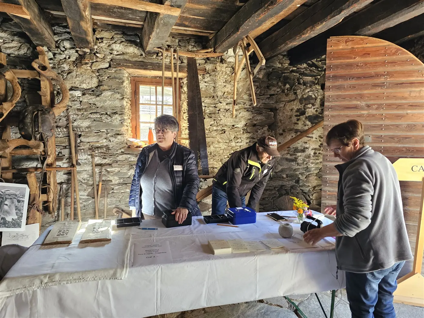 Tre persone stanno lavorando a un tavolo in una vecchia struttura di pietra, circondate da attrezzi rustici. Una persona è in piedi a sinistra, mentre le altre due sono concentrate su documenti e materiali sul tavolo. La luce entra da una finestra, creando un'atmosfera calda e accogliente.