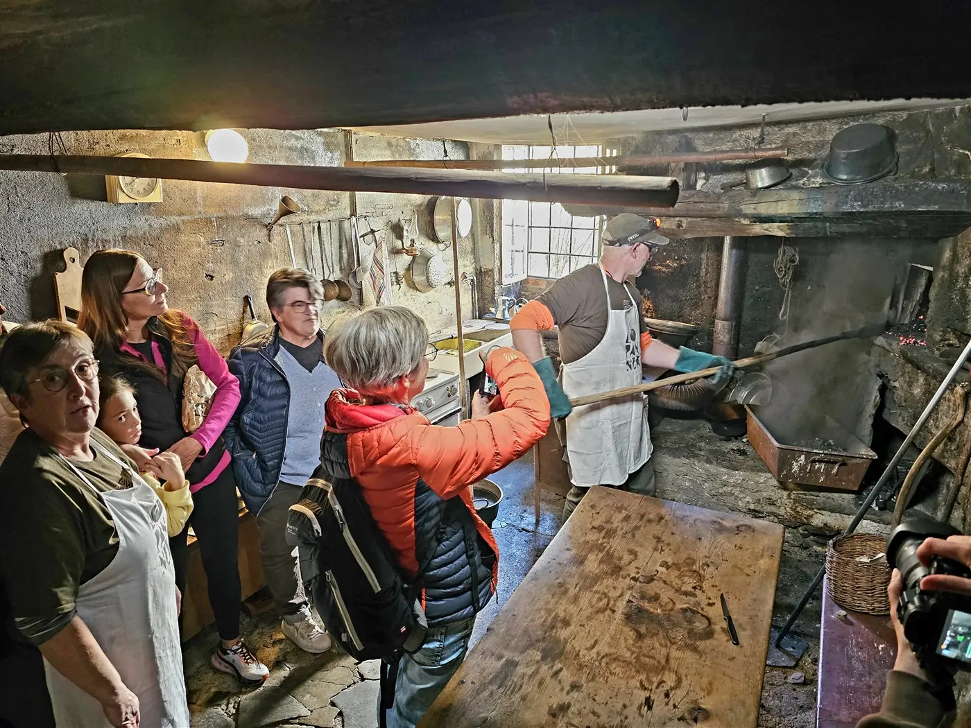 Un gruppo di persone osserva un cuoco mentre lavora in una cucina tradizionale. Sono presenti utensili da cucina appesi e un focolare. L'atmosfera è rustica e calda.