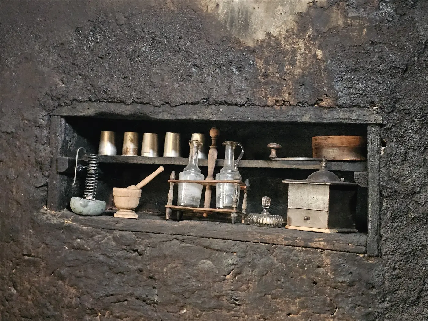 Ripiano in muratura con vari contenitori in vetro e metallo, tra cui bottiglie, un mortaio e un macinino, tutti disposti in un ambiente scuro e rustico.