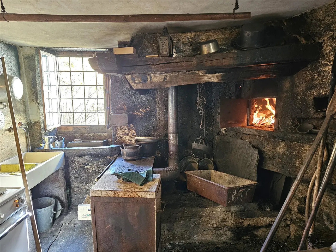 Cucina rustica con pareti in pietra e un camino acceso. Un lavello e un piano cottura sono visibili, mentre oggetti e utensili da cucina sono sparsi sul tavolo e intorno. La luce naturale entra da una finestra grande.