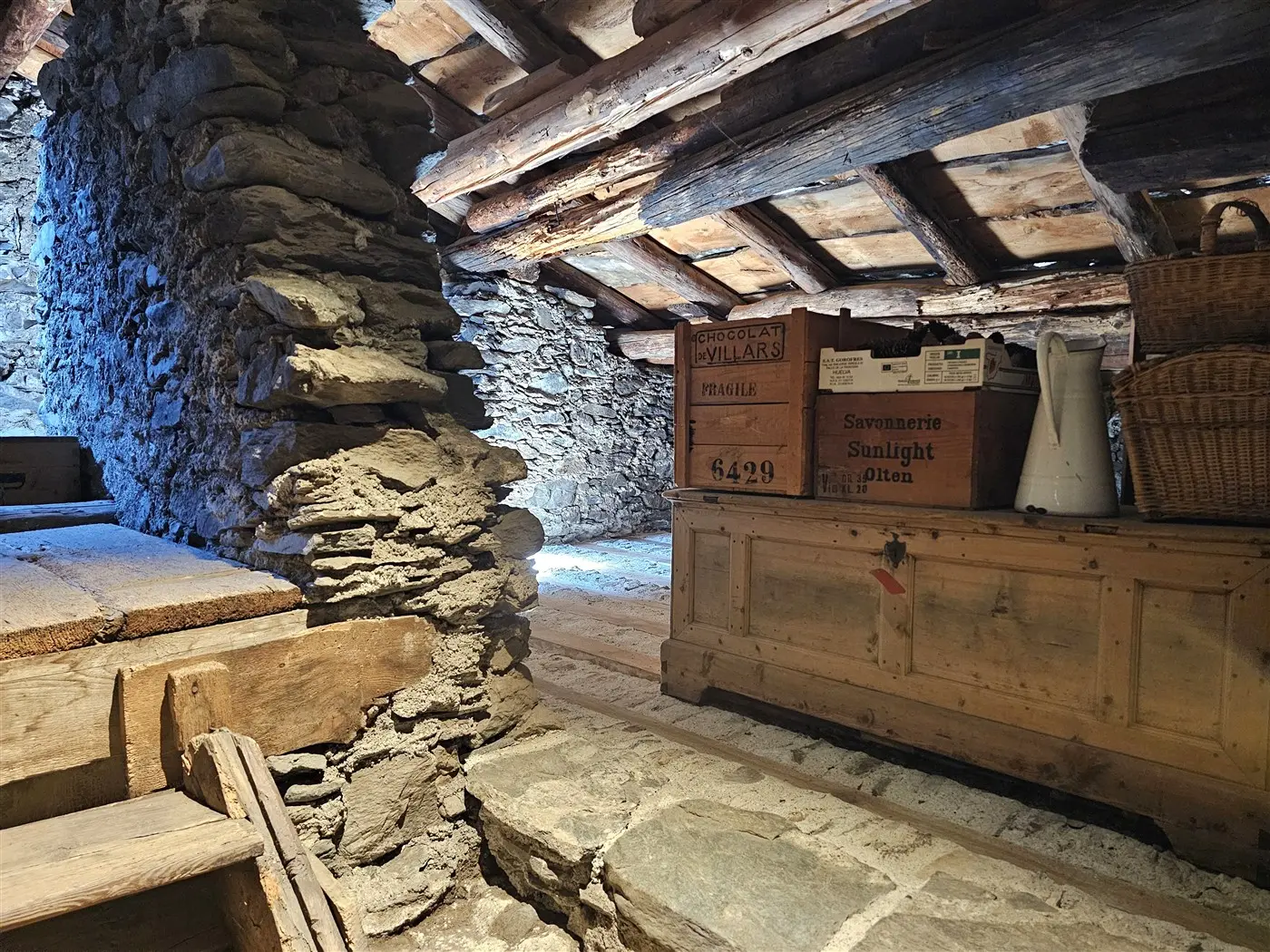 Interno di un rustico con pareti in pietra e soffitto in legno. Una scala di legno conduce verso un piano inferiore. Un banco in legno è visibile, adornato da ceste e contenitori. La luce naturale illumina l'ambiente, creando un'atmosfera calda e accogliente.