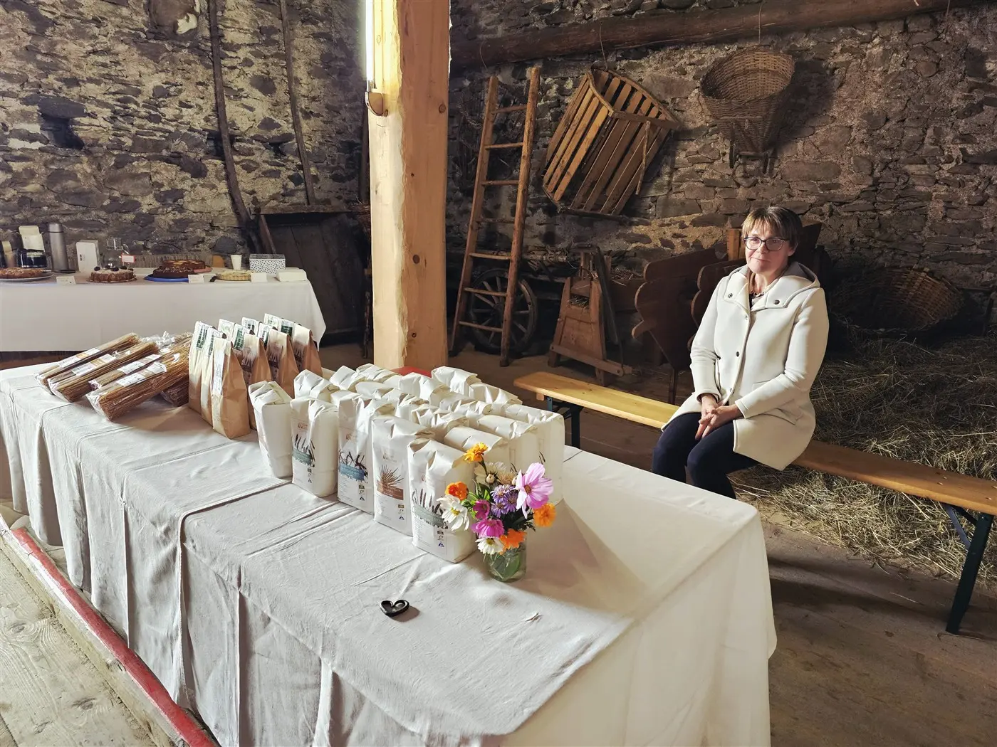 Una donna seduta su una panchina in un ambiente rustico, circondata da sacchetti di prodotti su un tavolo. Il tavolo è coperto da una tovaglia bianca e ci sono fiori in un vaso. Sullo sfondo, elementi di legno e attrezzature agricole.