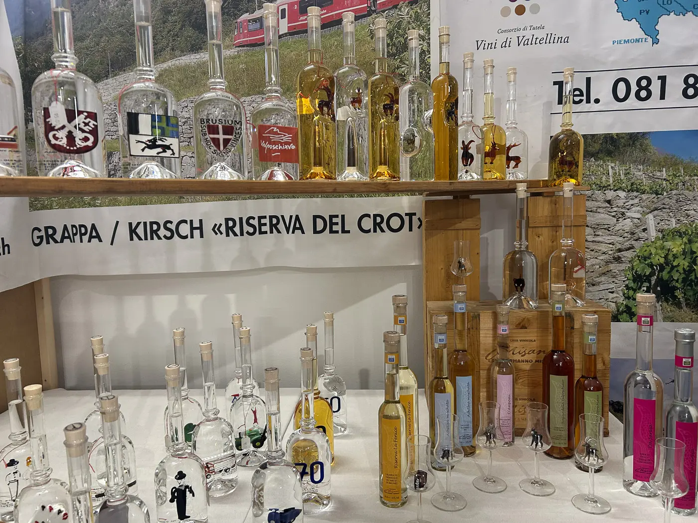 Una esposizione di bottiglie di grappa e kirsch, con etichette artistiche e vari colori, disposte su una tavola di legno. Sullo sfondo, un poster con informazioni sui vini della Valtellina.
