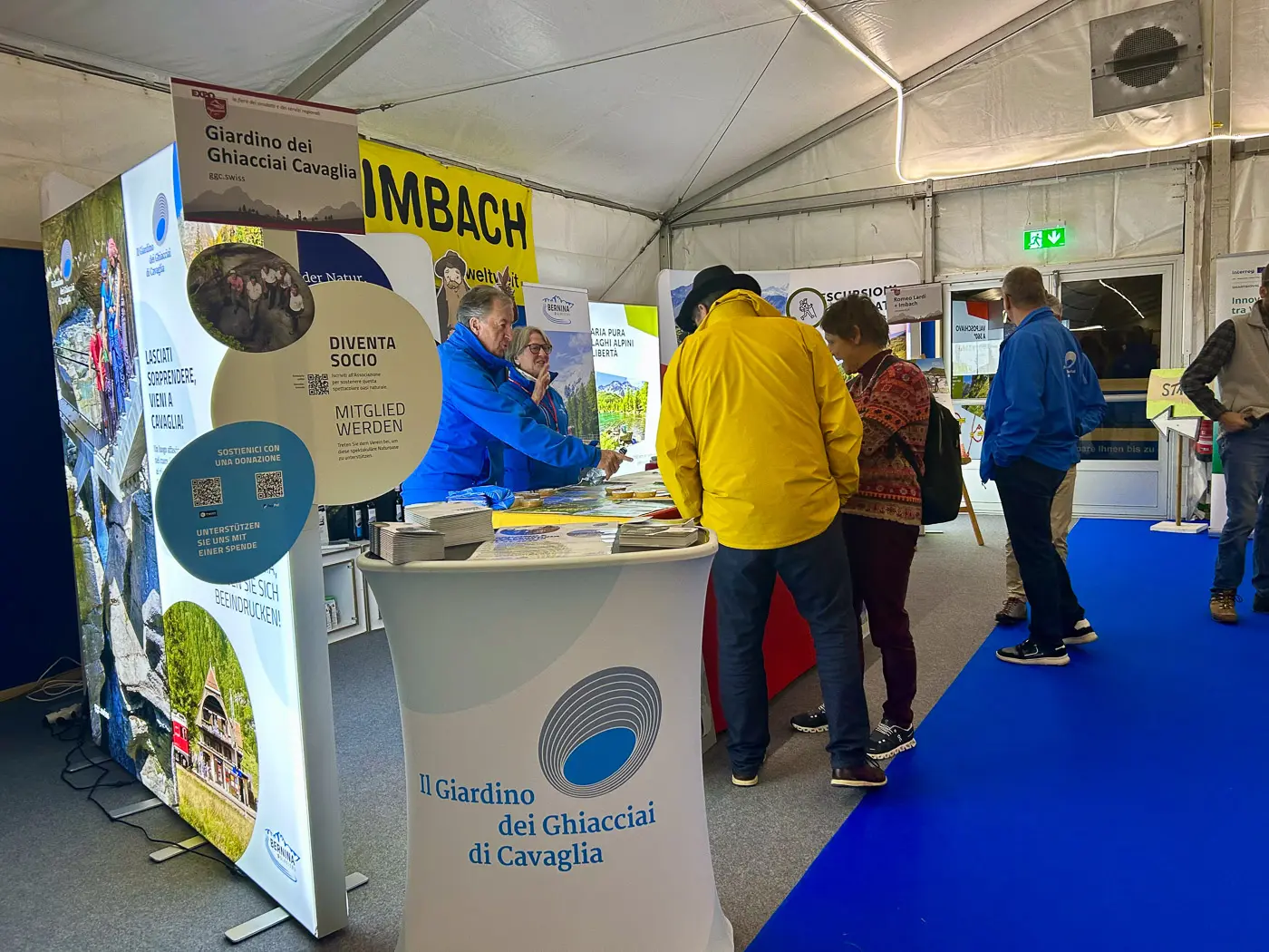 Uno stand espositivo con due pannelli informativi su "Il Giardino dei Ghiacciai di Cavaglia". Persone in interazione intorno a un tavolo con brochure e materiali promozionali. Ambiente interno con tende e pavimento blu.