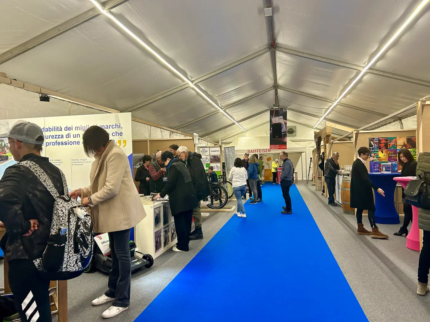 Un'area espositiva all'interno di una tenda. Persone interagiscono con vari stand, alcuni osservano prodotti, altri chiacchierano. Il pavimento è ricoperto da un tappeto blu.