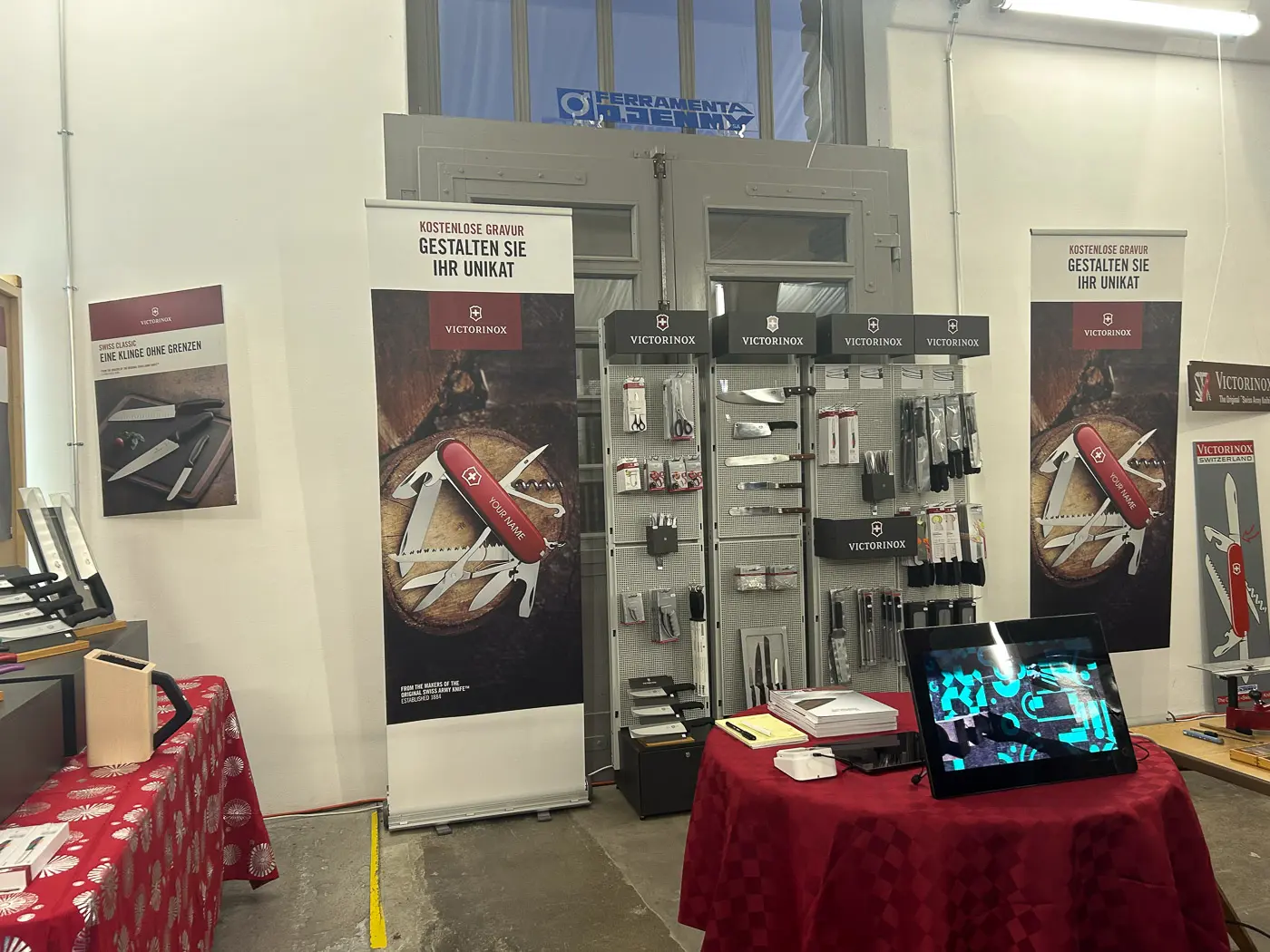 Espazio espositivo con banner e prodotti Victorinox. Sono visibili diversi coltelli e attrezzi esposti su una parete, con un tavolo centrale ricoperto da un tessuto rosso. Schermo visibile con presentazione di prodotti.