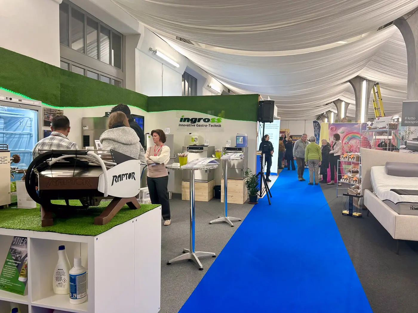 Una fiera espositiva con stand allestiti e visitatori che interagiscono. I colori predominanti sono il verde e il blu, con diverse aziende che presentano i loro prodotti e innovazioni.