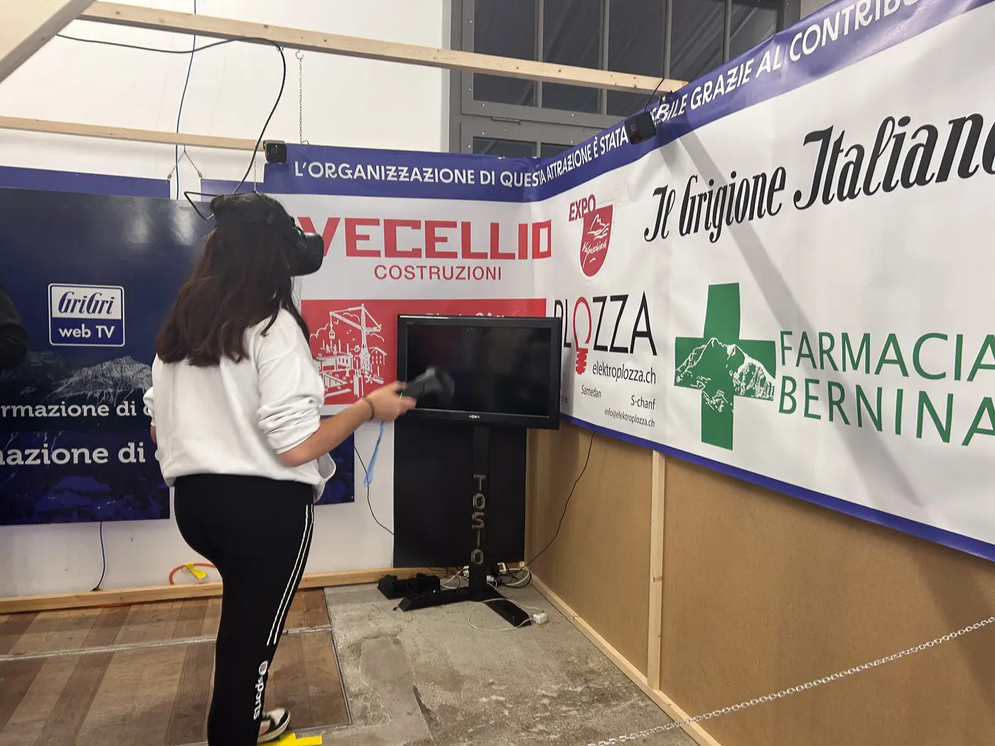 Una persona utilizza un visore di realtà virtuale all'interno di uno stand espositivo, circondata da banchetti e cartelloni pubblicitari.