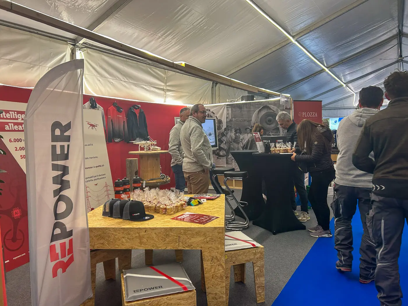 Stand espositivo con un bancone e un tavolo in legno. Sul tavolo ci sono bicchieri di vino e prodotti in vista. Sullo sfondo si vedono varie attrezzature e un banner del marchio "REPPOWER". Persone che discutono e si muov