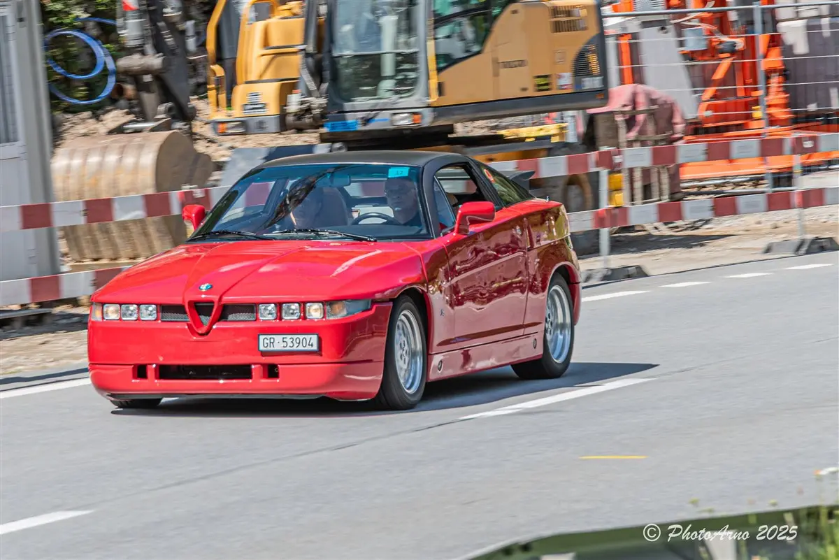 Una sportiva rossa Alfa Romeo si muove lungo una strada in un ambiente urbano. Sullo sfondo, macchinari da cantiere e barriere di sicurezza sono visibili.