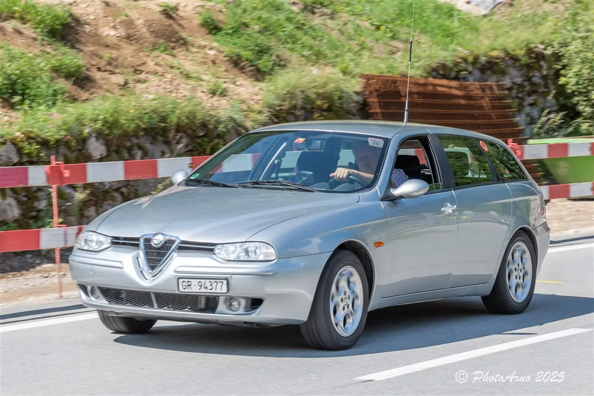 Un'auto berlina argentata sta viaggiando su una strada. Il veicolo è un'Alfa Romeo, con targa visibile, e in lontananza si notano barriere di contenimento. Le montagne fanno da sfondo, suggerendo un ambiente naturale.