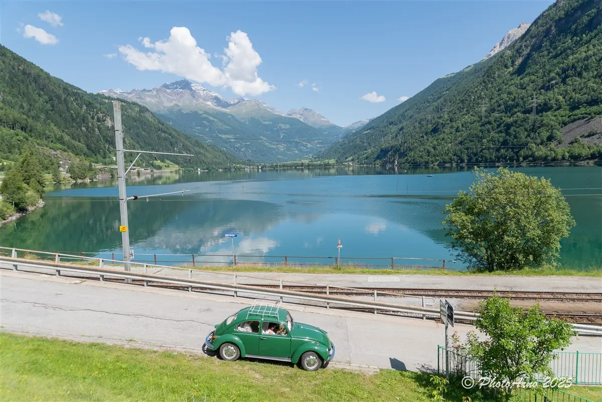 Un'auto verde vintage parcheggiata lungo una strada, con un lago riflettente e montagne sullo sfondo. Il cielo è sereno e parzialmente nuvoloso.