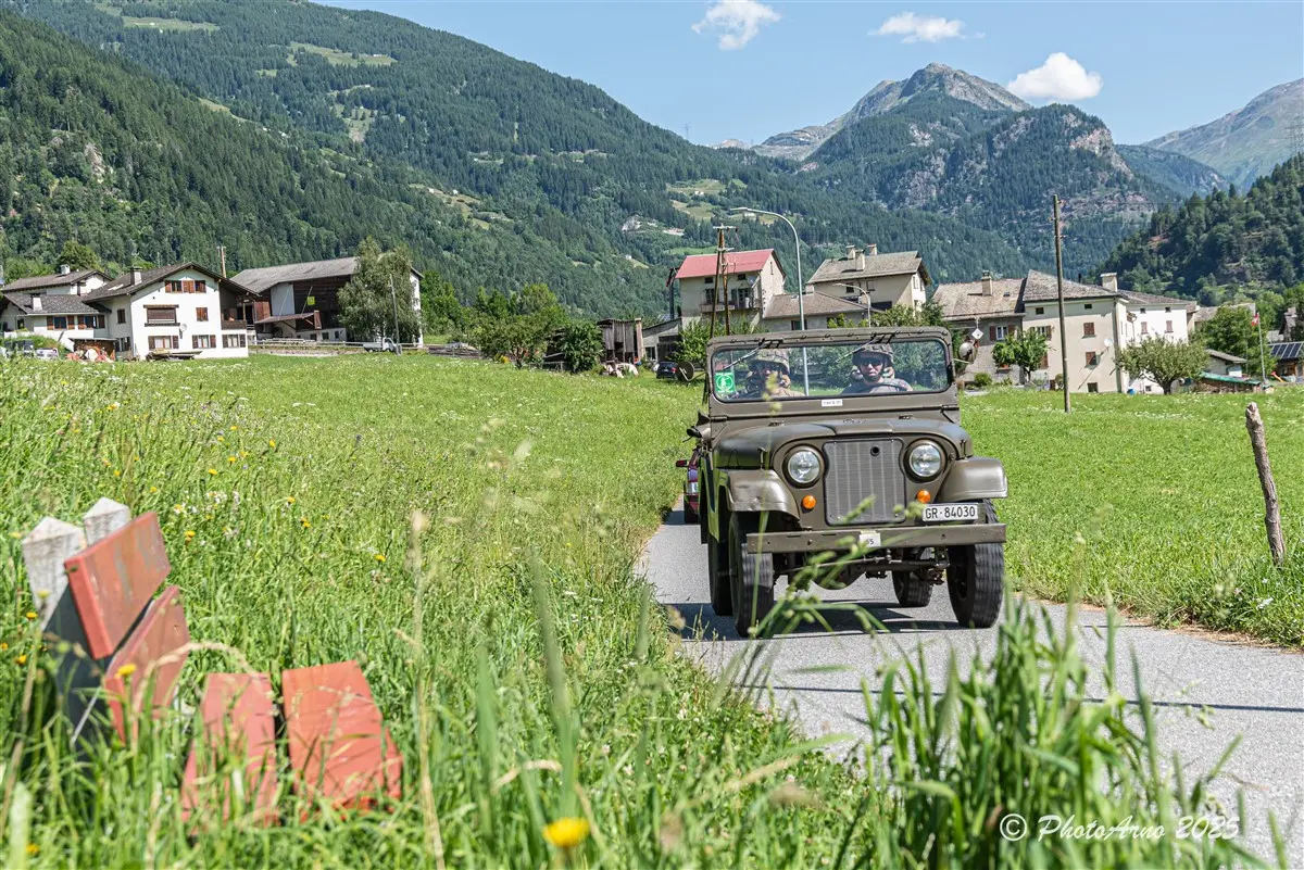 Un'auto militare vintage percorre una strada panoramica circondata da verdi prati e montagne. Si possono vedere edifici di legno e case di montagna sullo sfondo. Il cielo è sereno e soleggiato.