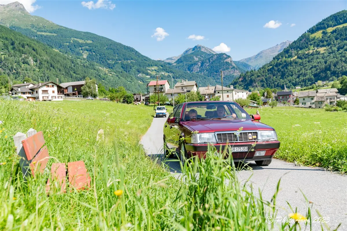 Un'auto rossa percorre una strada di campagna circondata da prati verdi e montagne. Sullo sfondo si intravedono alcune case e un altro veicolo. Il cielo è sereno e soleggiato.